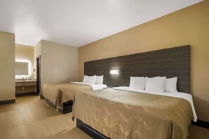 A hotel room with two beds, a large dark wood headboard, a bathroom with a mirror and sink, and a light-colored floor.