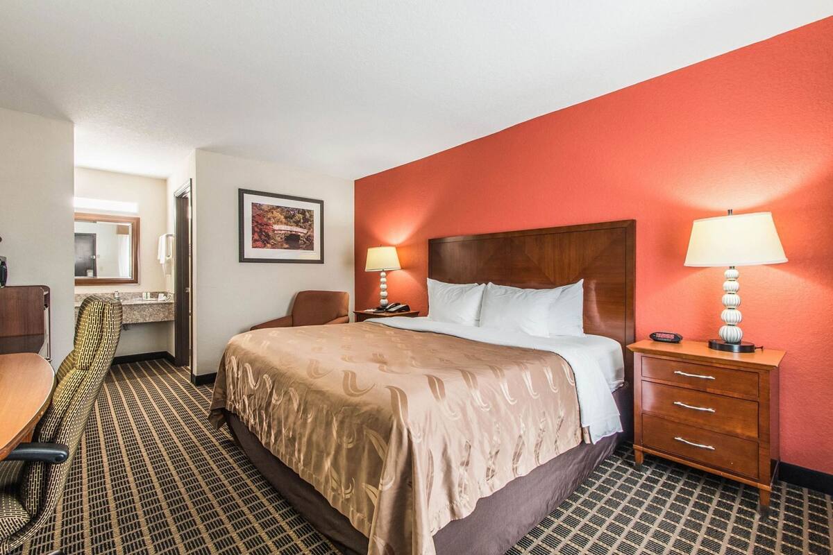 A hotel room with a bed, a desk, a chair, a nightstand with a lamp, and a bathroom visible in the background.