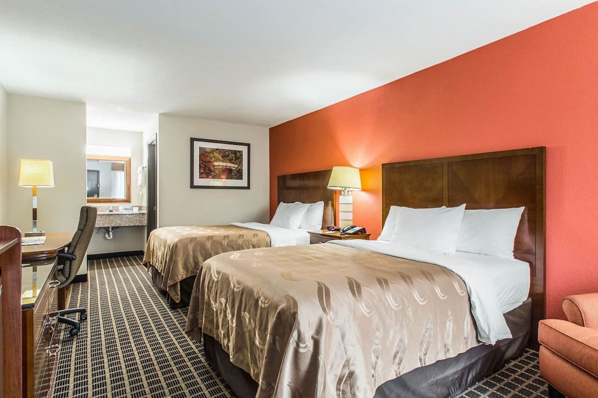 A hotel room with two beds, a desk, a chair, a lamp, and a painting on the wall.