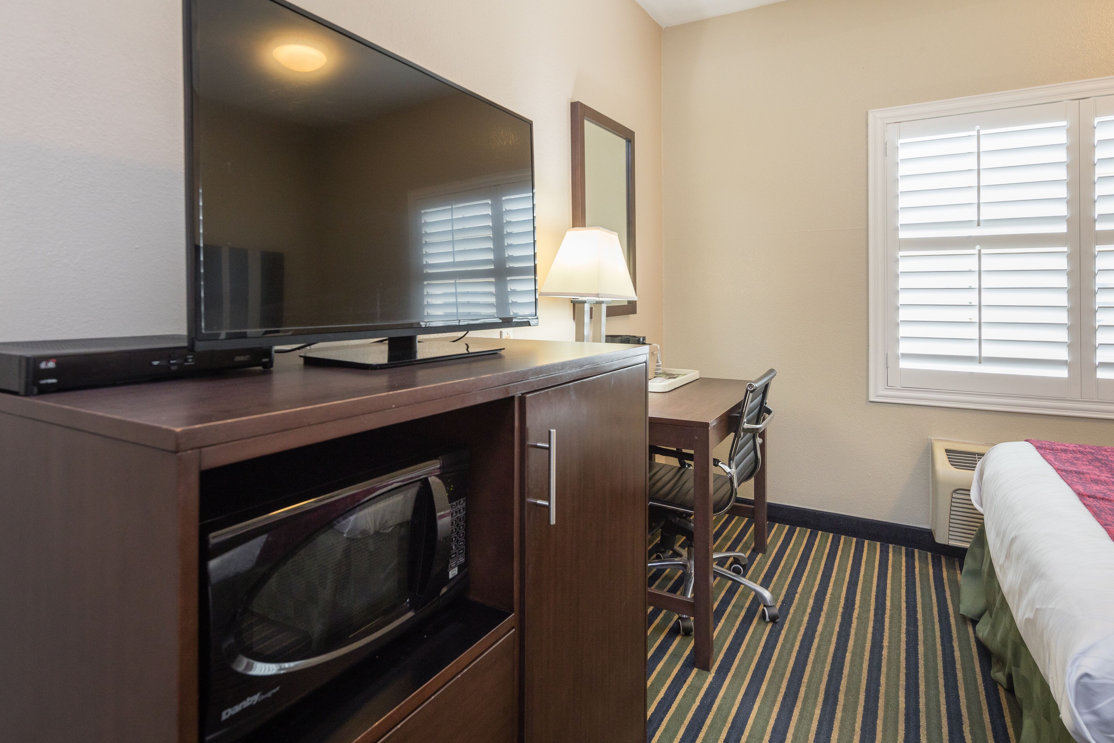 A hotel room with a flat-screen TV, a desk with a lamp, a microwave, a bed, and a window with blinds.