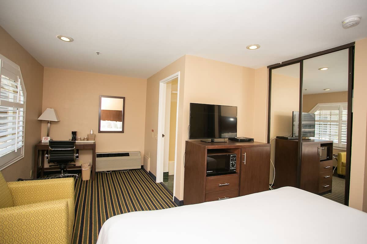 A hotel room with a bed, a desk with a chair, a television, a mirror, and a window with blinds.