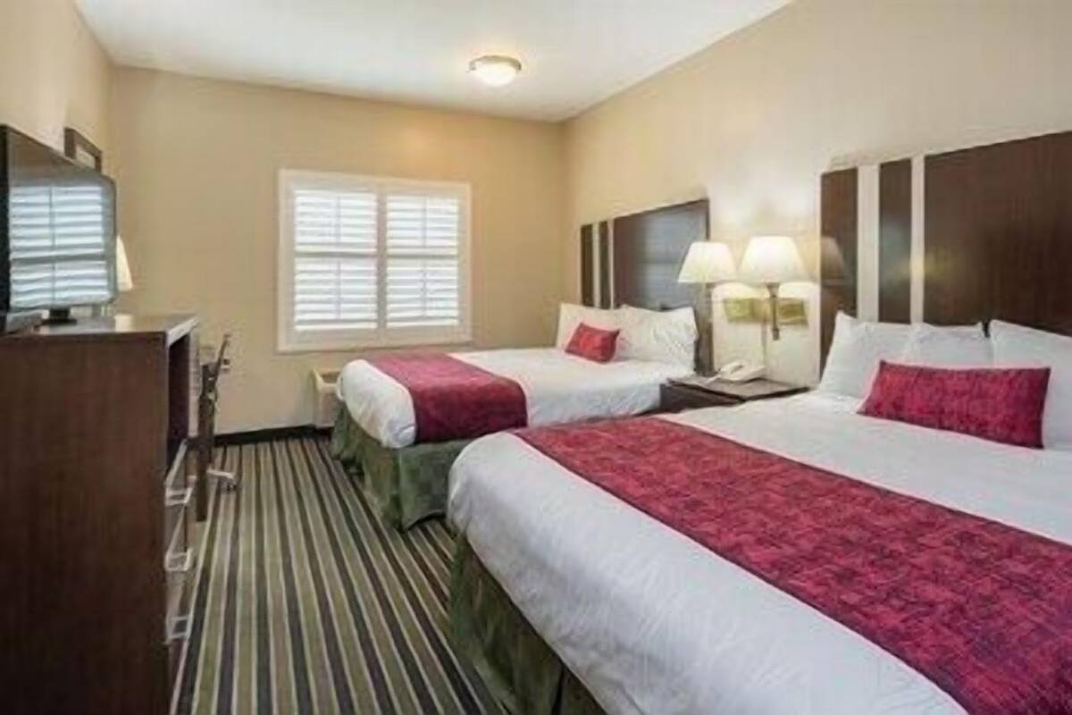 A hotel room with two beds, a desk, a lamp, and a window.