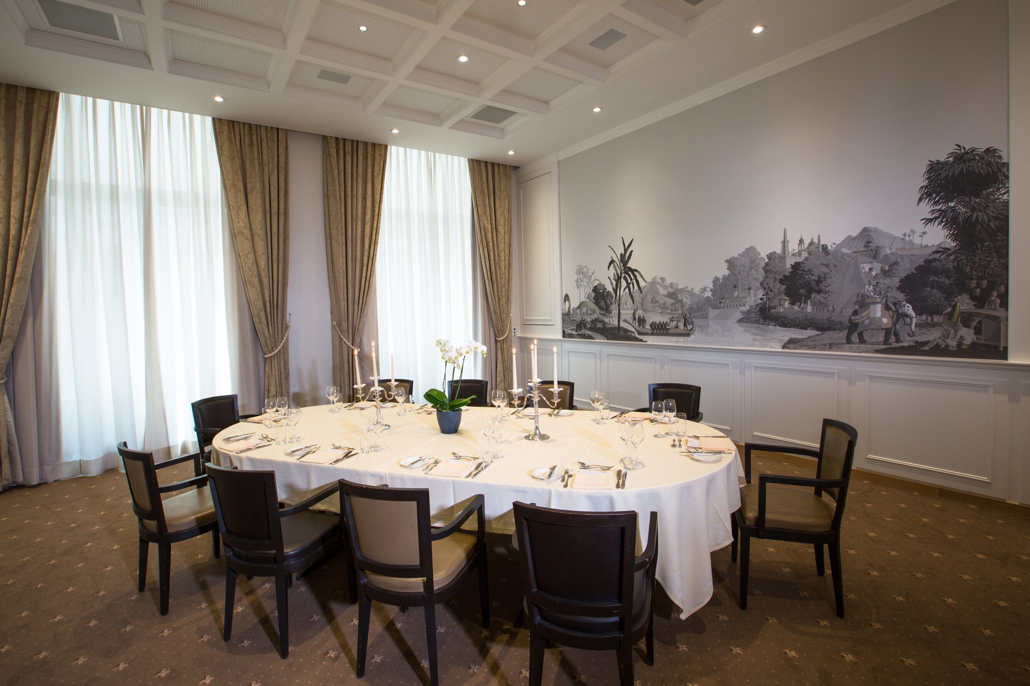 A dining room with a round table set for six, surrounded by chairs, with a large wall mural depicting a landscape scene.