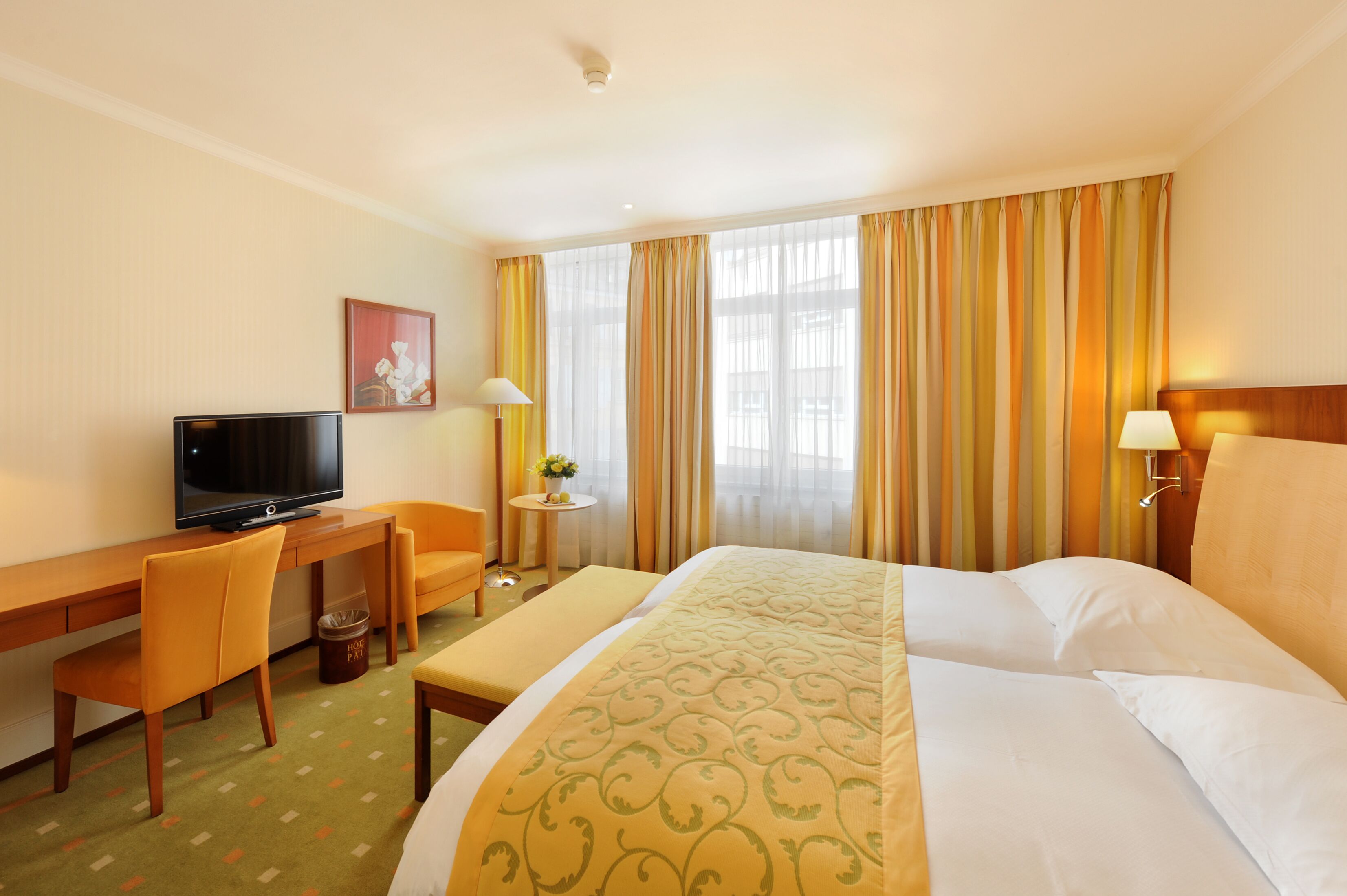 Habitación de hotel con cama, escritorio con televisor, silla, mesita de noche con lámpara y ventana con cortinas.