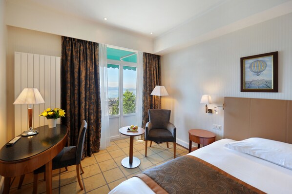 Habitación de hotel con cama, escritorio con lámpara, silla, una pequeña mesa con frutas y vistas al exterior.