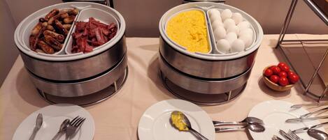 Disposição de buffet com duas grandes travessas de aço inoxidável, uma com salsichas e presunto, e outra com ovos mexidos e ovos cozidos.