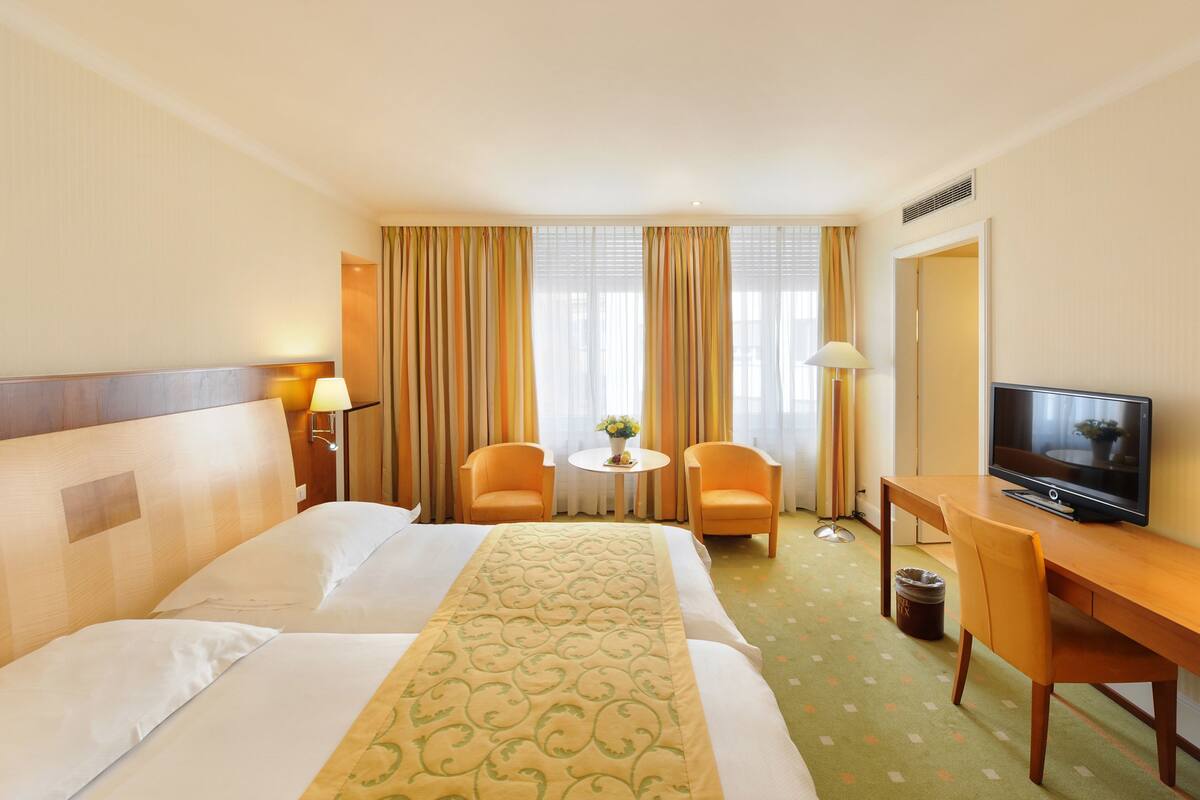 A hotel room with a bed, two chairs, a small table with a vase, a desk with a television, and a window with curtains.