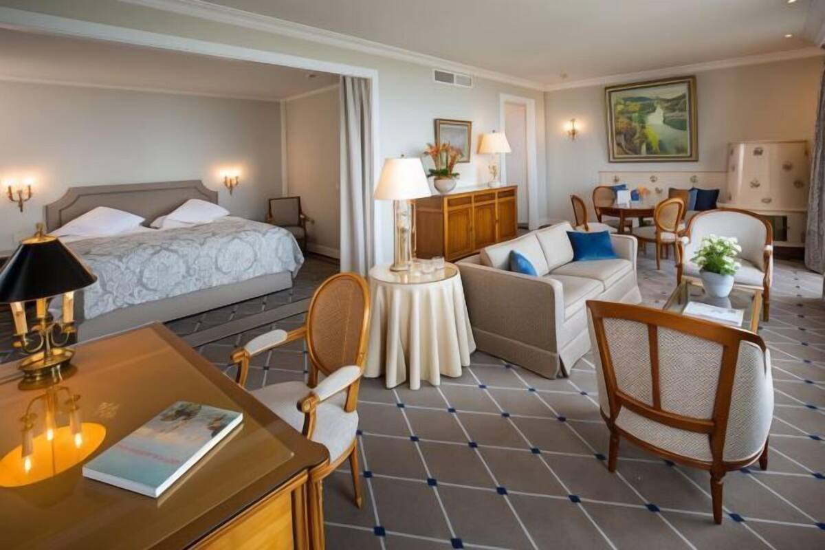 A hotel room with a bed, a sofa, a dining table with chairs, a small round table with a lamp, and a painting on the wall.