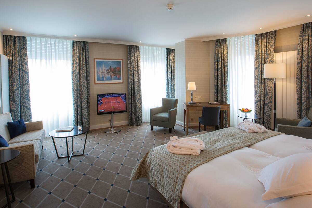 A hotel room with a bed, two armchairs, a desk, a TV, and a small table.