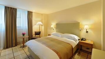 A hotel room with a large bed, two bedside tables, a small round table with a lamp, and a view of the outside through sheer curtains.