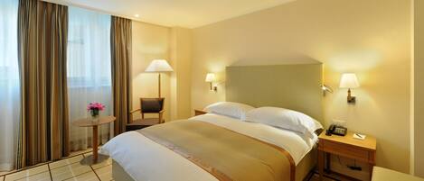 Habitación de hotel con una cama grande, dos mesitas de noche, una mesita redonda con lámpara y vistas al exterior a través de cortinas de tela ligera.