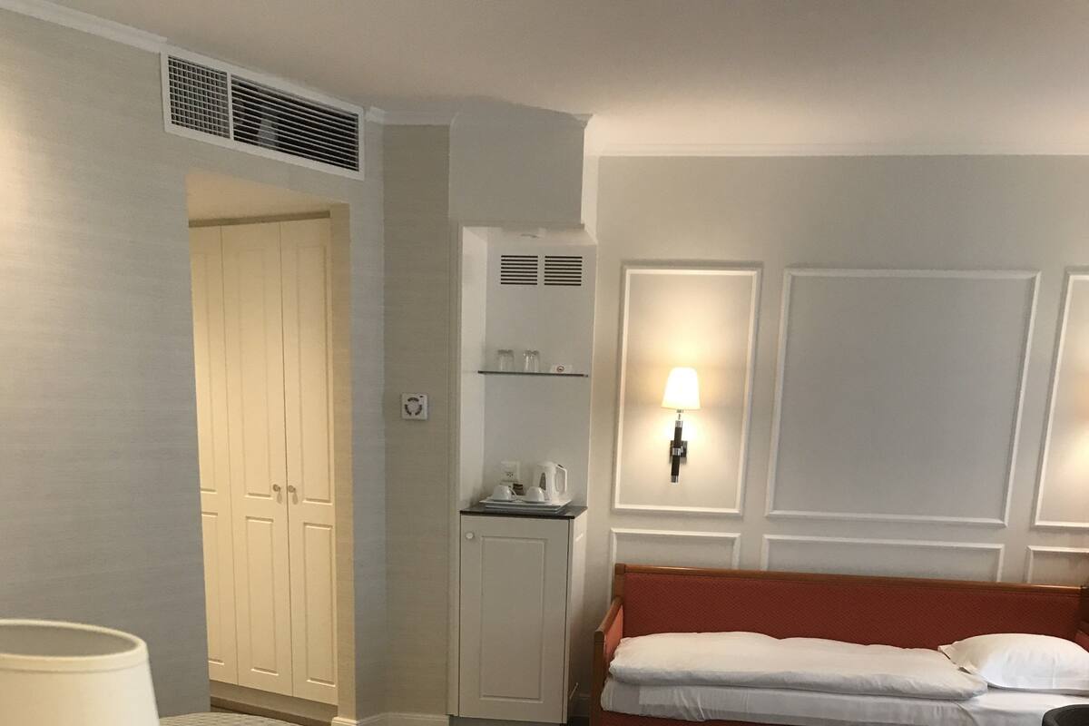 A hotel room with a bed, a bedside table with a lamp, a small cabinet, a wardrobe, and a wall-mounted heater.
