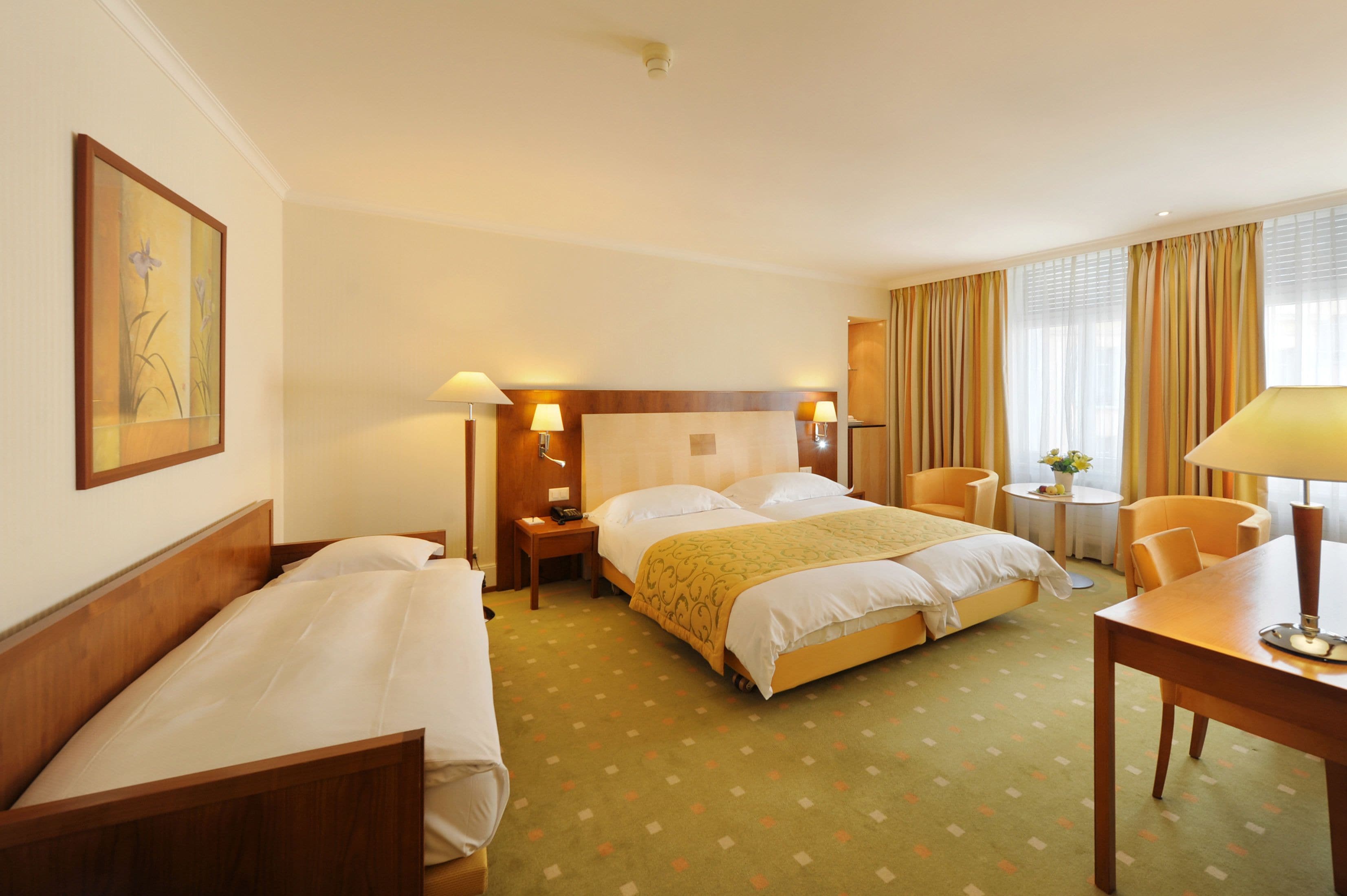 A hotel room with a large bed, two bedside tables with lamps, a small round table with a chair, and a painting on the wall.