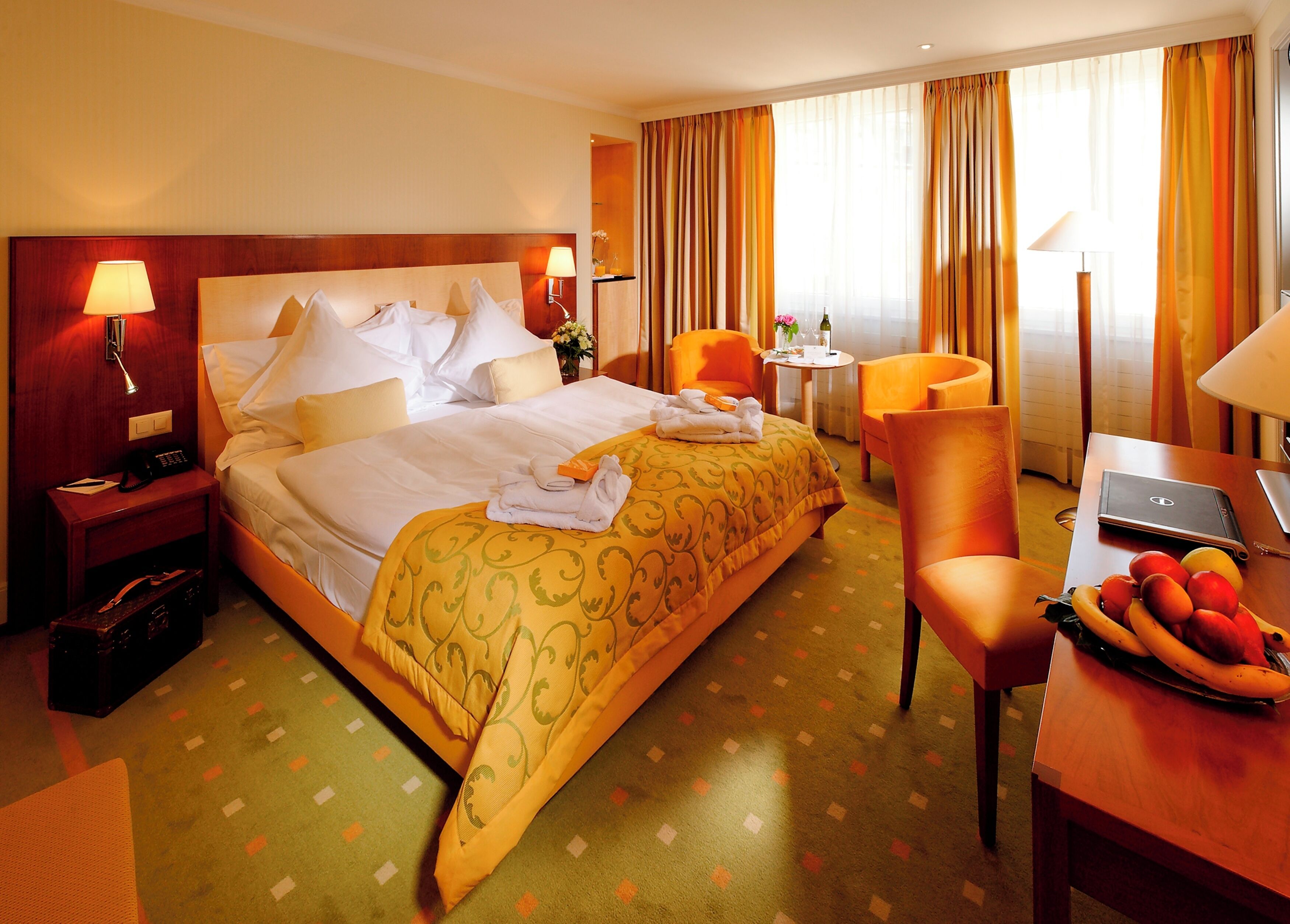 Une chambre d’hôtel avec un grand lit, un bureau avec un ordinateur portable, une chaise, une petite table avec un vase et un panier de fruits.