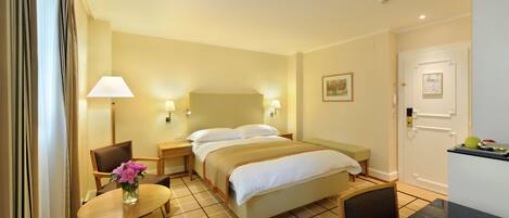 A hotel room with a bed, bedside tables with lamps, a small table with a vase of flowers, a chair, and a kitchenette area.