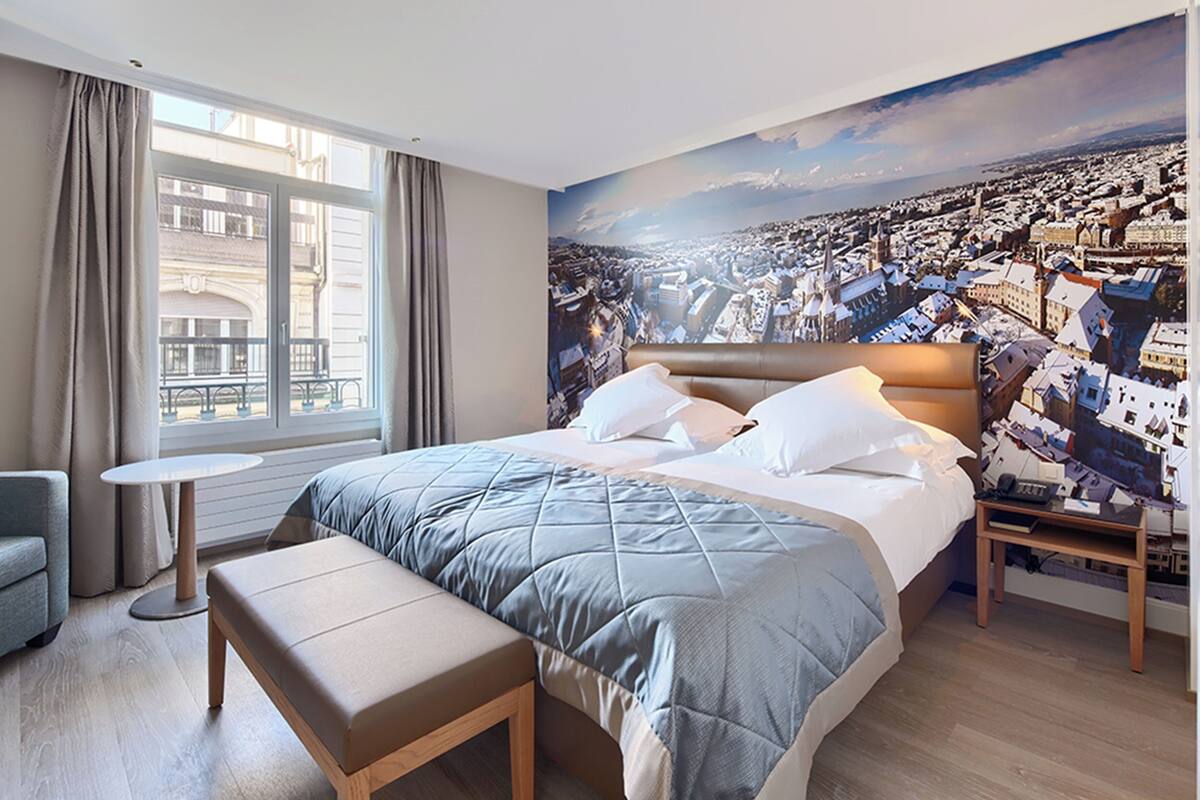 A hotel room with a large bed, a small round table, a bench, and a cityscape mural on the wall.