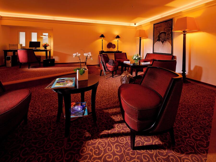 Ein Hotelzimmer mit rotem Teppich, einem Schreibtisch mit Computer, zwei Stühlen, einem Tisch mit Blumenvase und einer Lampe.