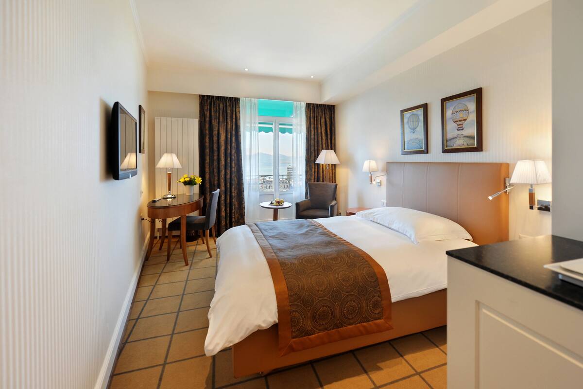 A hotel room with a bed, a desk, a chair, a small table, and a window with curtains.