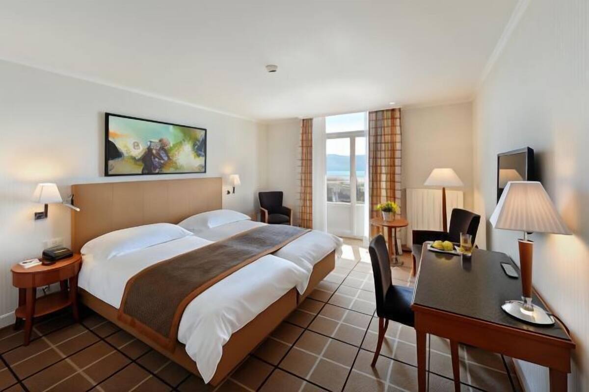 A hotel room with a bed, two bedside tables, a desk, chairs, a TV, and a window with curtains.