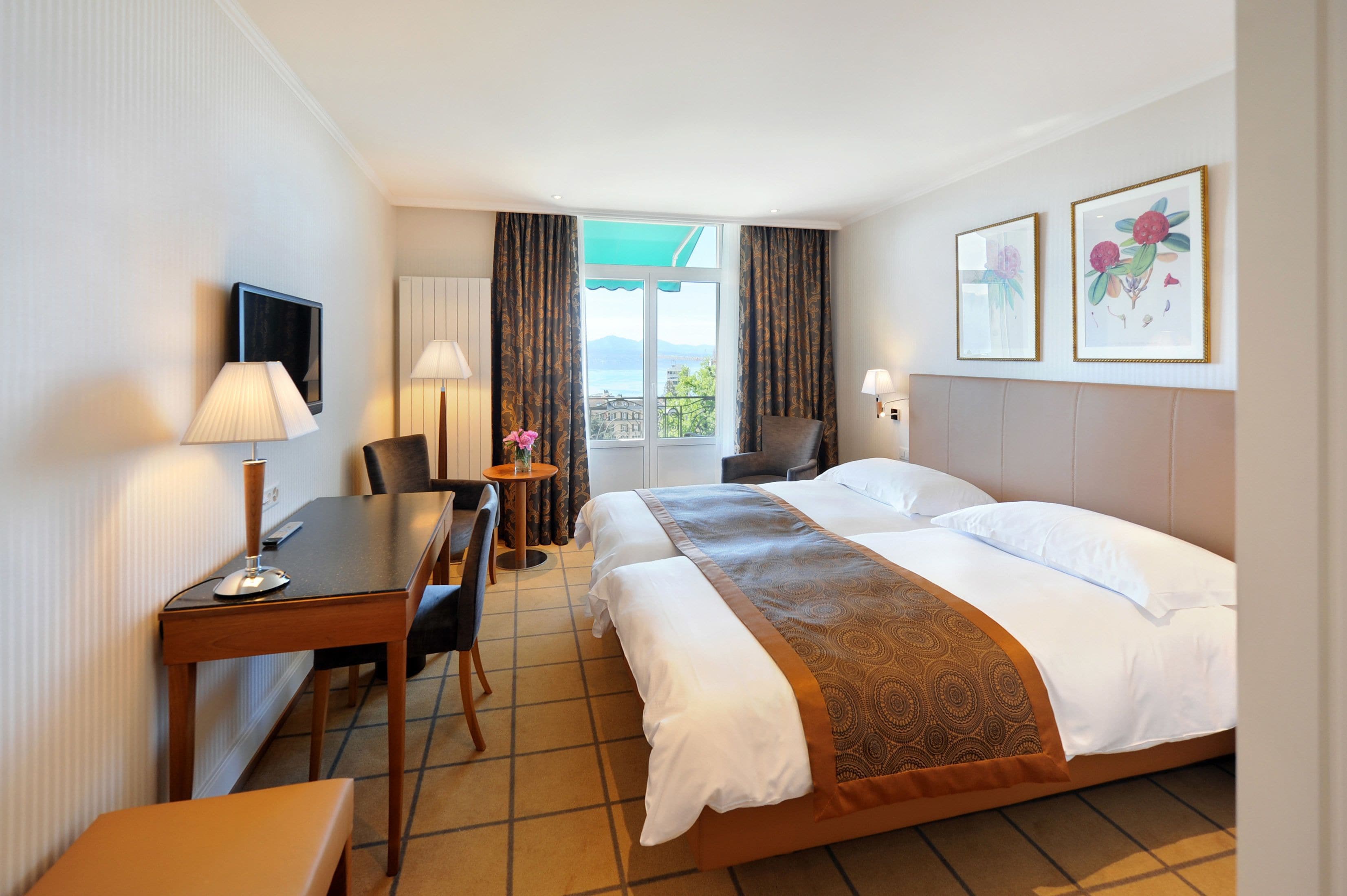 A hotel room with a bed, a desk, a chair, a small table with a vase, and two framed pictures on the wall.