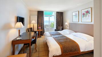 A hotel room with a bed, a desk, a chair, a small table with a vase, and two framed pictures on the wall.