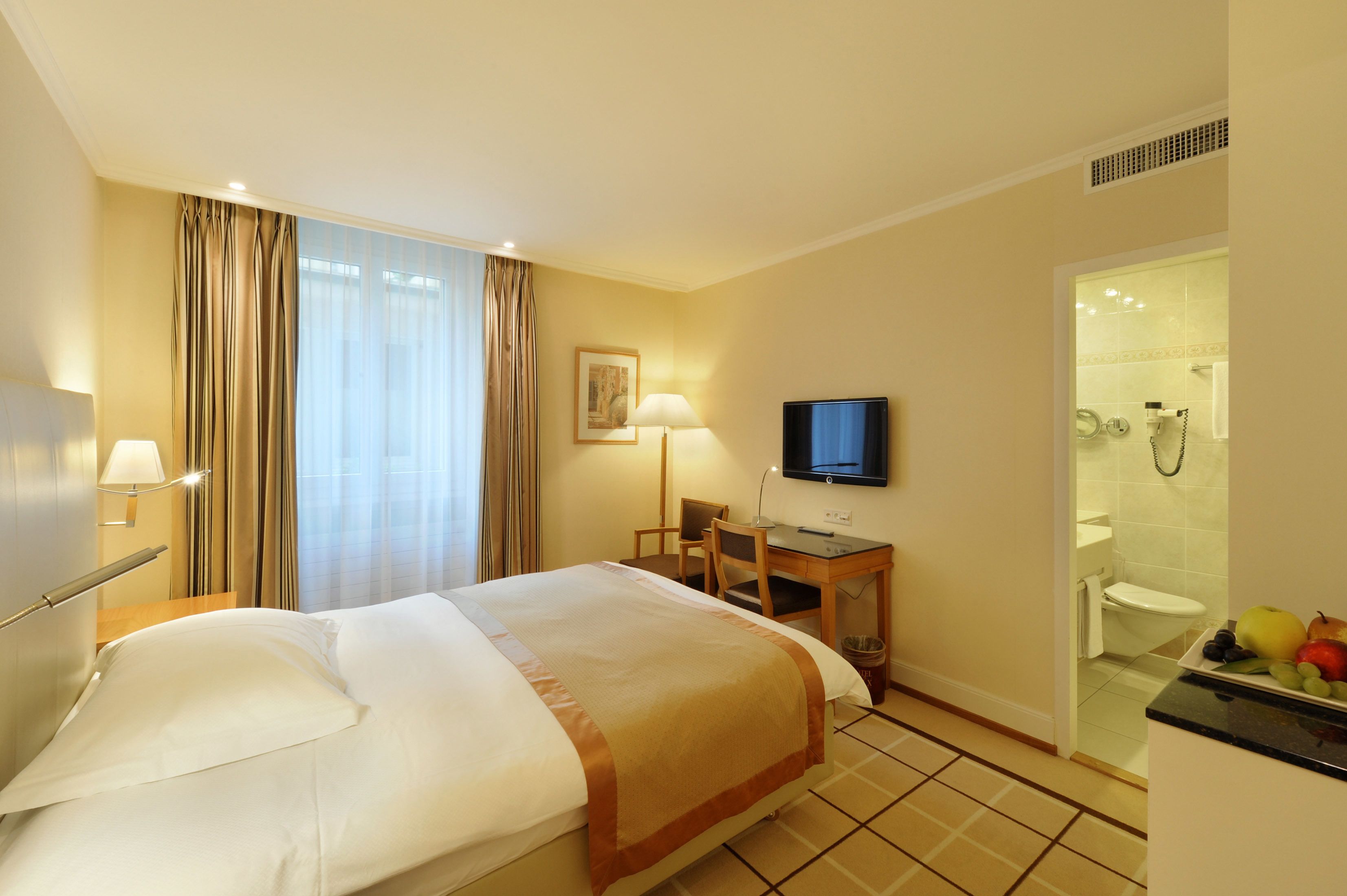Ein Hotelzimmer mit Bett, Schreibtisch, Stuhl, Fernseher und einem durch eine Glastür sichtbaren Badezimmer.