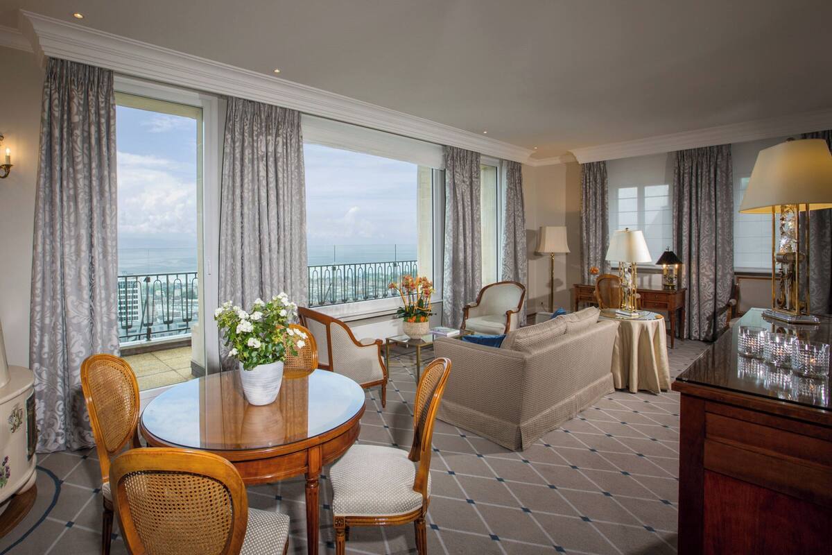 A hotel room with a balcony, a round dining table, wicker chairs, a sofa, and a side table with a lamp.