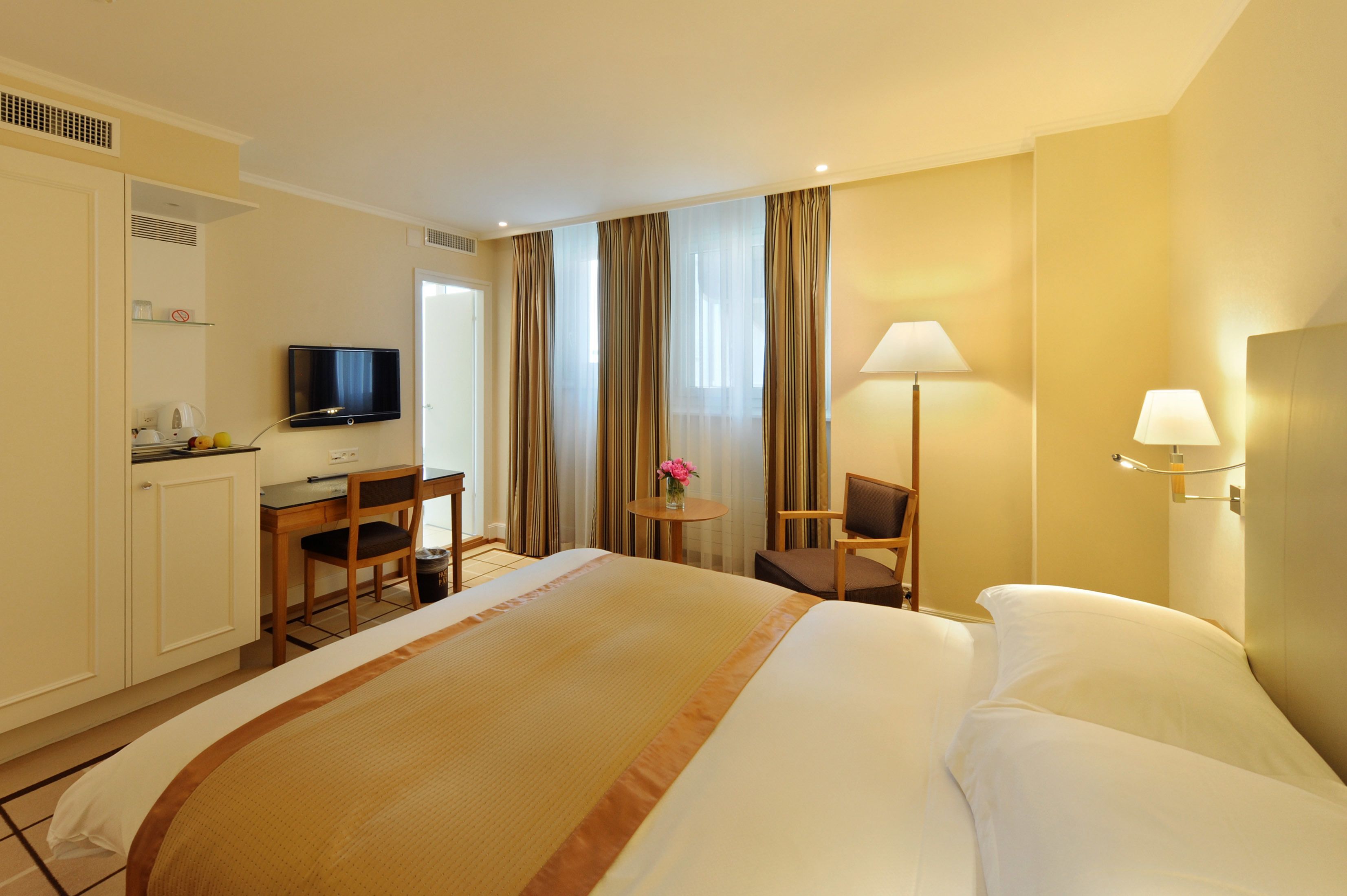 A hotel room with a large bed, a desk with a chair, a television, a lamp, and a small table with a vase of flowers.