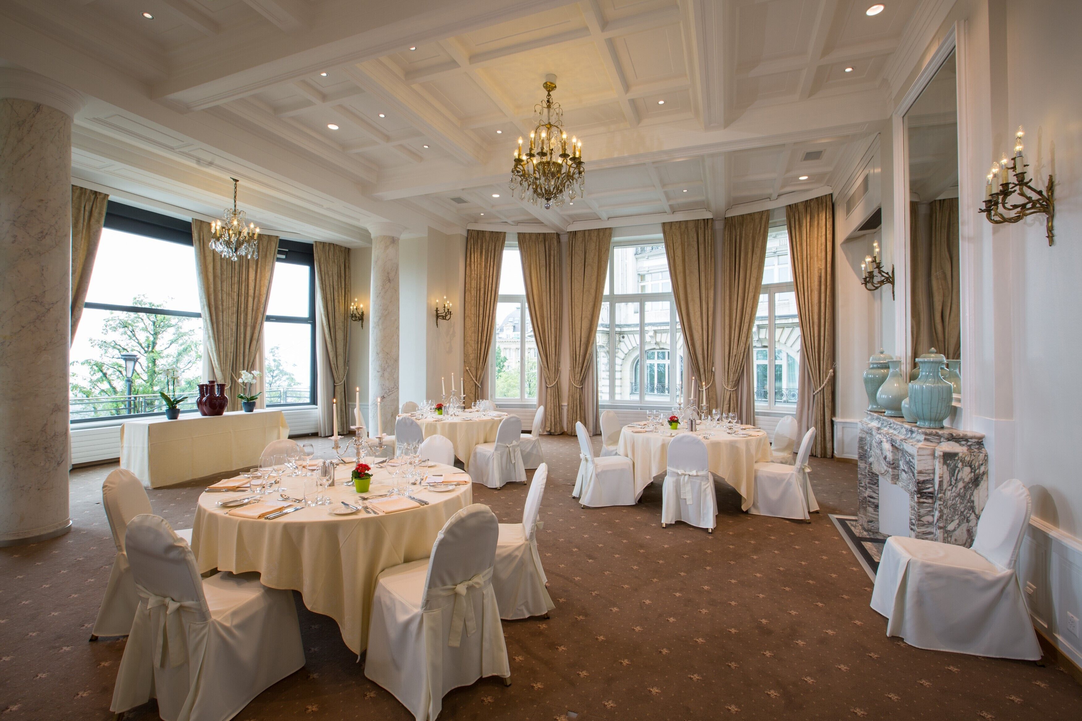 A formal dining room with round tables, white chairs, chandeliers, and large windows with curtains.