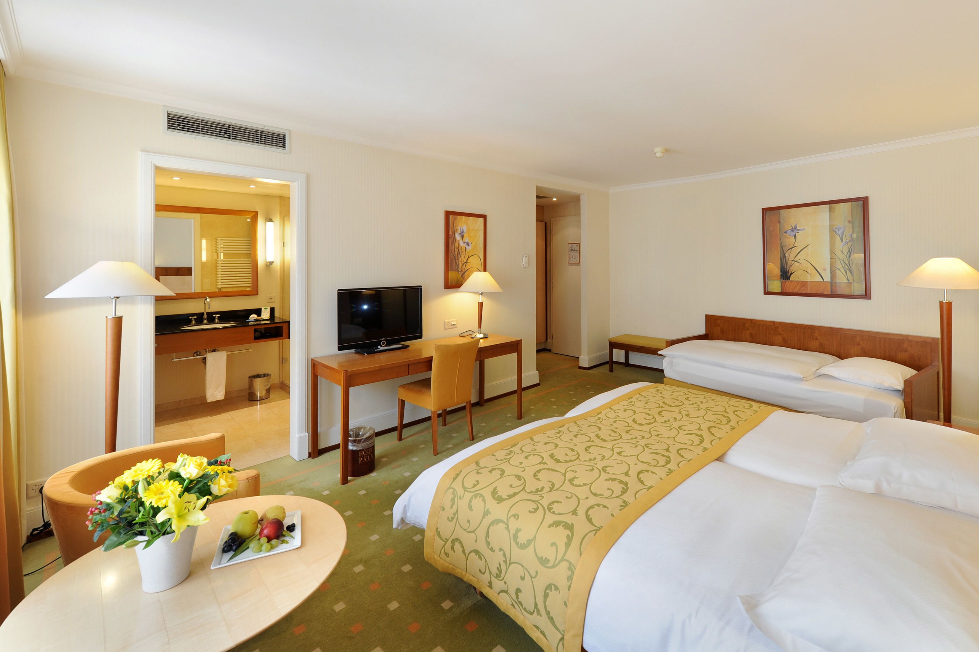 A hotel room with a bed, a desk with a computer, a TV, a bathroom, and a dining area with flowers and fruit.