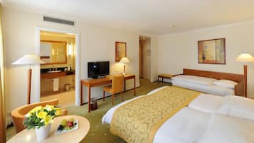 A hotel room with a bed, a desk with a computer, a TV, a bathroom, and a dining area with flowers and fruit.
