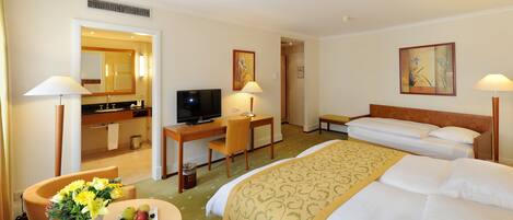A hotel room with a bed, a desk with a computer, a TV, a bathroom, and a dining area with flowers and fruit.
