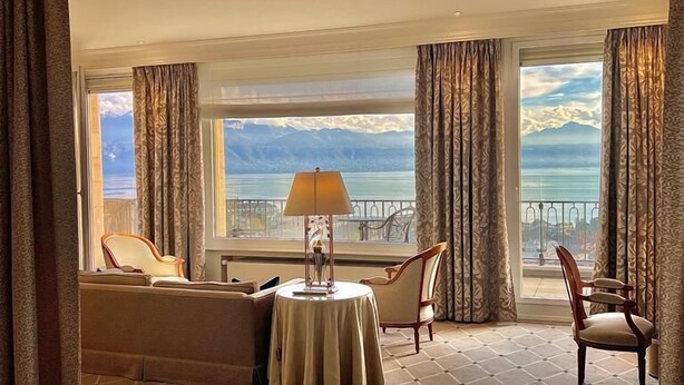 A hotel room with a bed, a small round table with a lamp, two chairs, and a view of a mountainous landscape through large windows. - De la Paix (Lausanne)