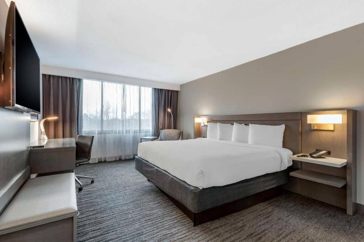 Une chambre d’hôtel avec un grand lit, un bureau, une chaise, une télévision, une table de chevet et une fenêtre avec des rideaux.