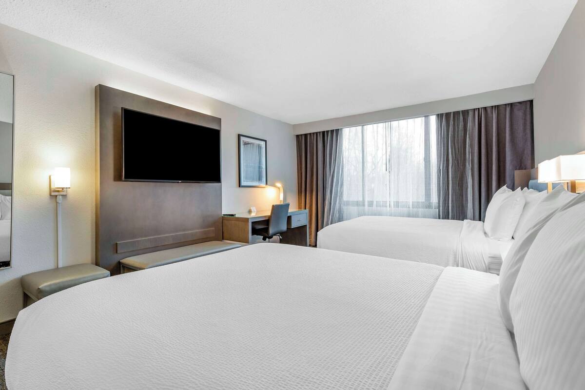 Une chambre d’hôtel avec deux lits, un bureau, une chaise, une télévision et une fenêtre avec des rideaux.