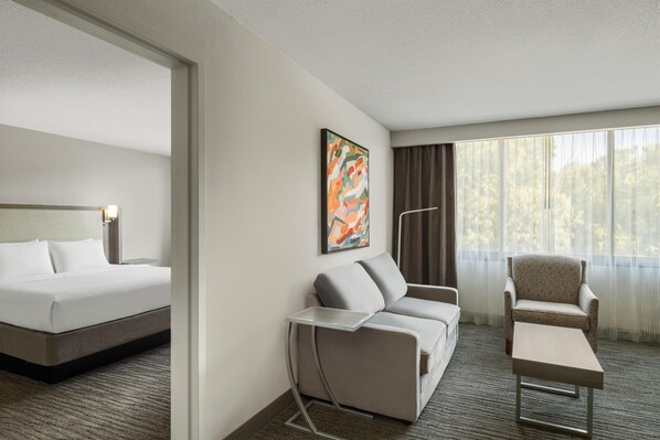 Una habitación de hotel con cama, sofá, sillón, una mesita y ventana con cortinas.