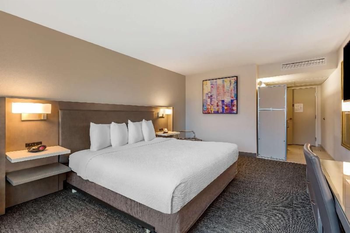 A hotel room with a bed, bedside tables, a TV, a painting on the wall, and a door leading to another room.