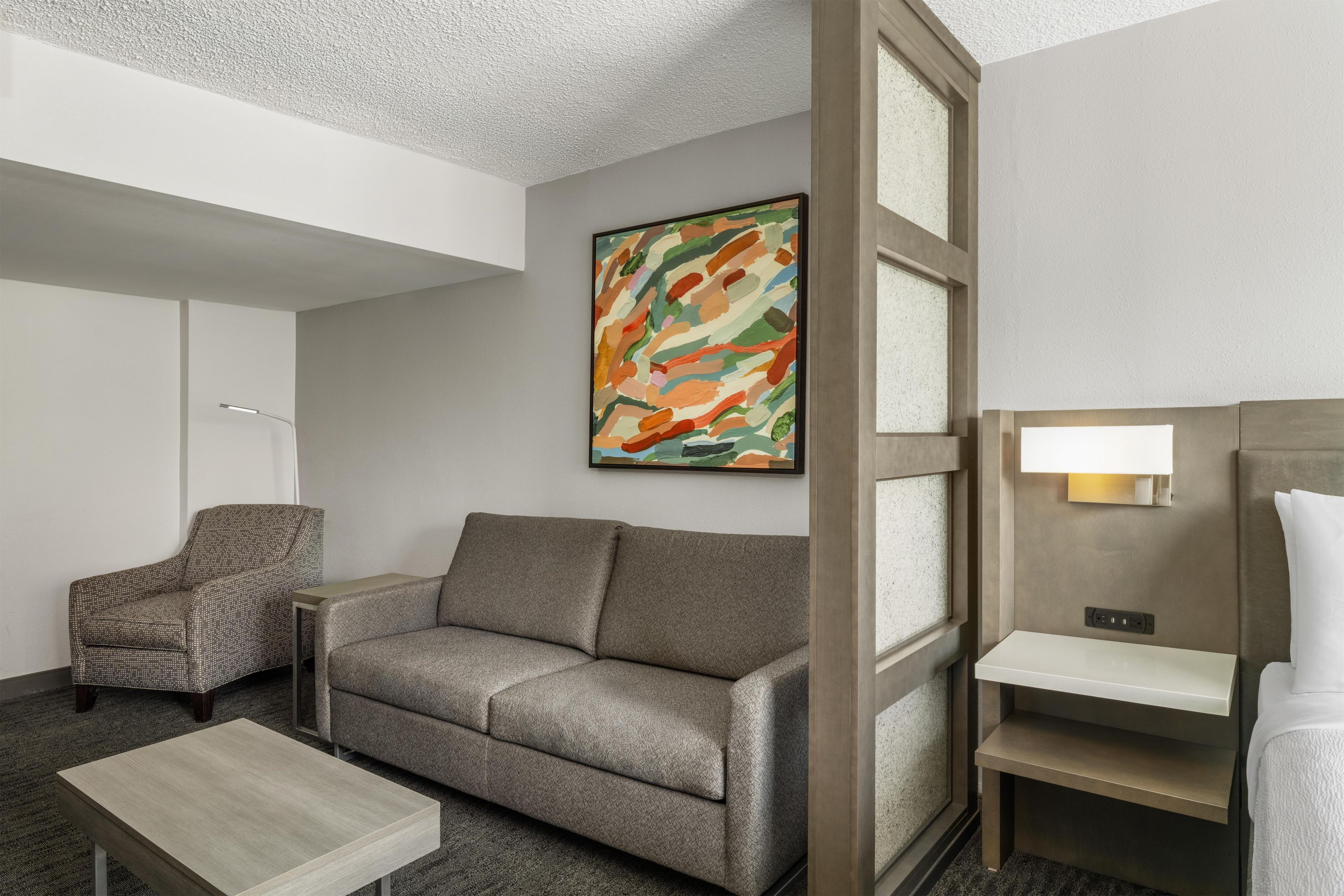 A hotel room with a grey sofa, a small table, a chair, a painting, and a bedside table with a lamp.