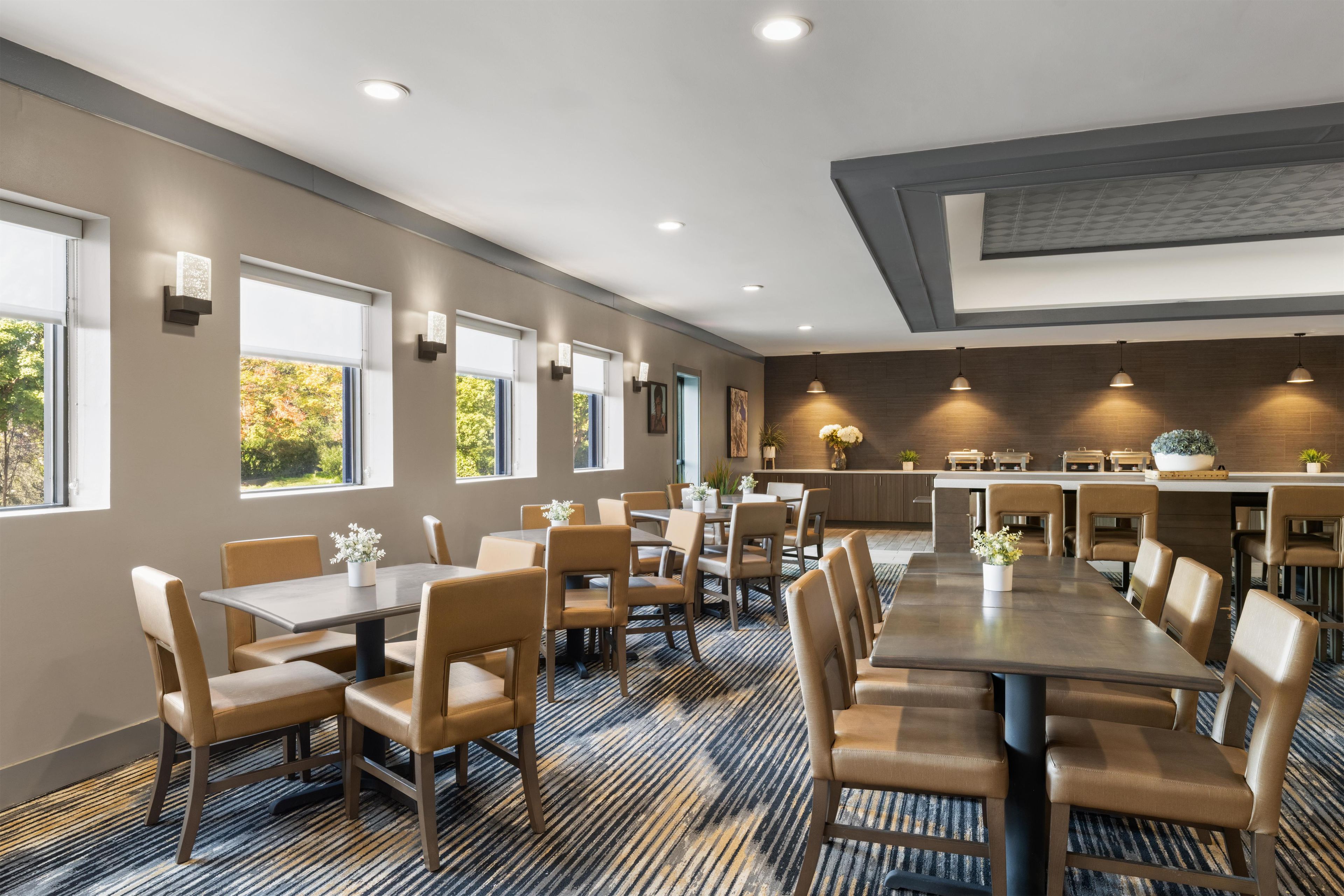Un comedor de hotel con varias mesas y sillas, una cocina al fondo y amplios ventanales que dejan entrar luz natural.