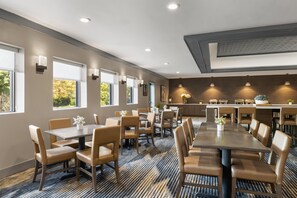 Un comedor de hotel con varias mesas y sillas, una cocina al fondo y amplios ventanales que dejan entrar luz natural.