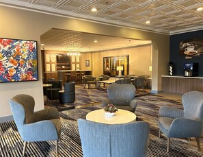 Eine Hotelhalle im modernen Design, mit einer Sitzecke mit mehreren Stühlen und Tischen, einer Bar und einem großen abstrakten Gemälde an der Wand.