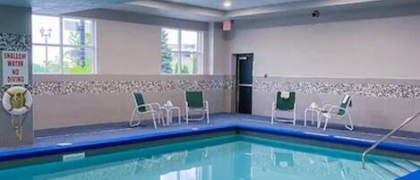 Une piscine intérieure à l’eau d’un bleu cristallin, entourée de chaises blanches et bordée d’un mur carrelé sur lequel est apposé un panneau d’avertissement.
