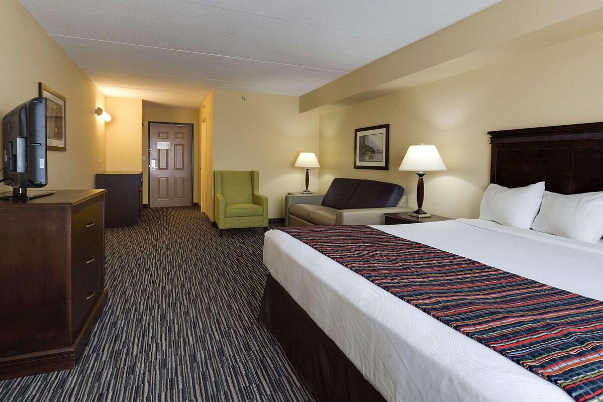 A hotel room with a bed, a desk with a computer, a chair, a lamp, and a door.