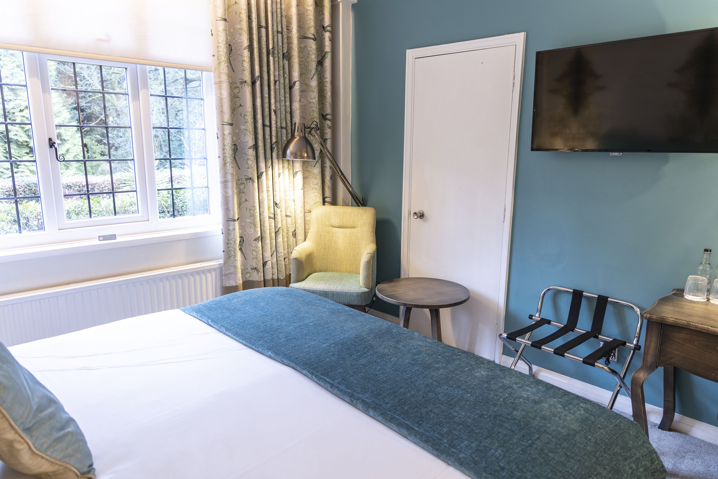 A bedroom with a bed, a chair, a small table, a TV, and a window with curtains.