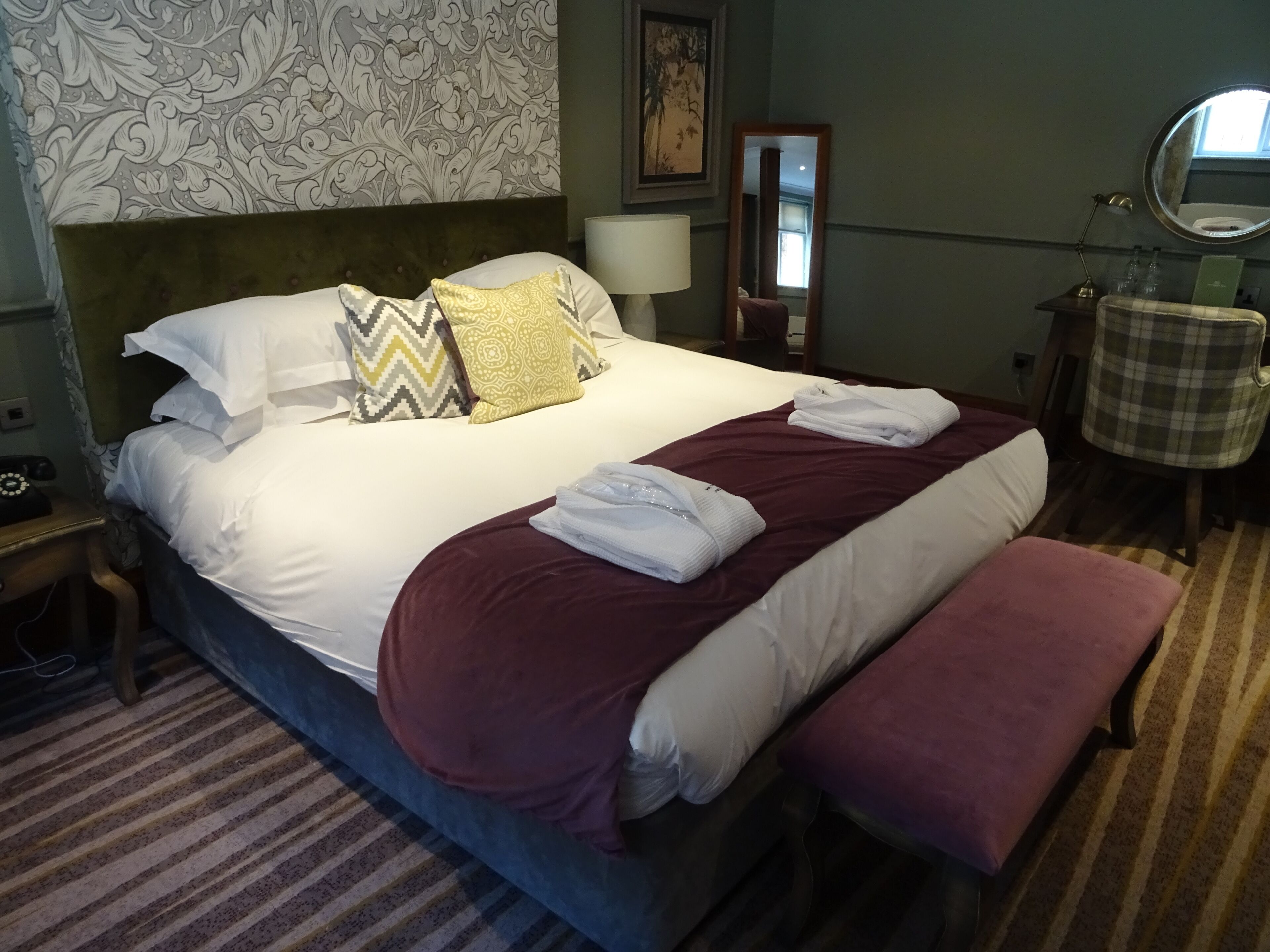 Una cama bien tendida con almohadas estampadas, una lámpara de noche, un espejo y una silla a cuadros.
