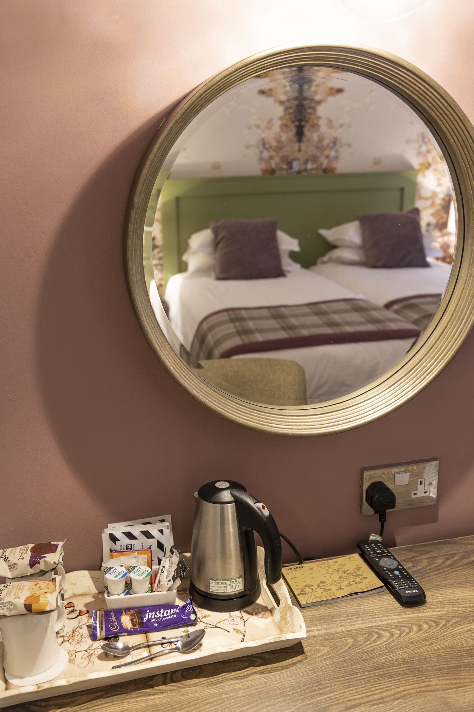 A hotel room with a bed, a bedside table with a kettle, a remote control, and a tray with snacks and a chocolate bar.
