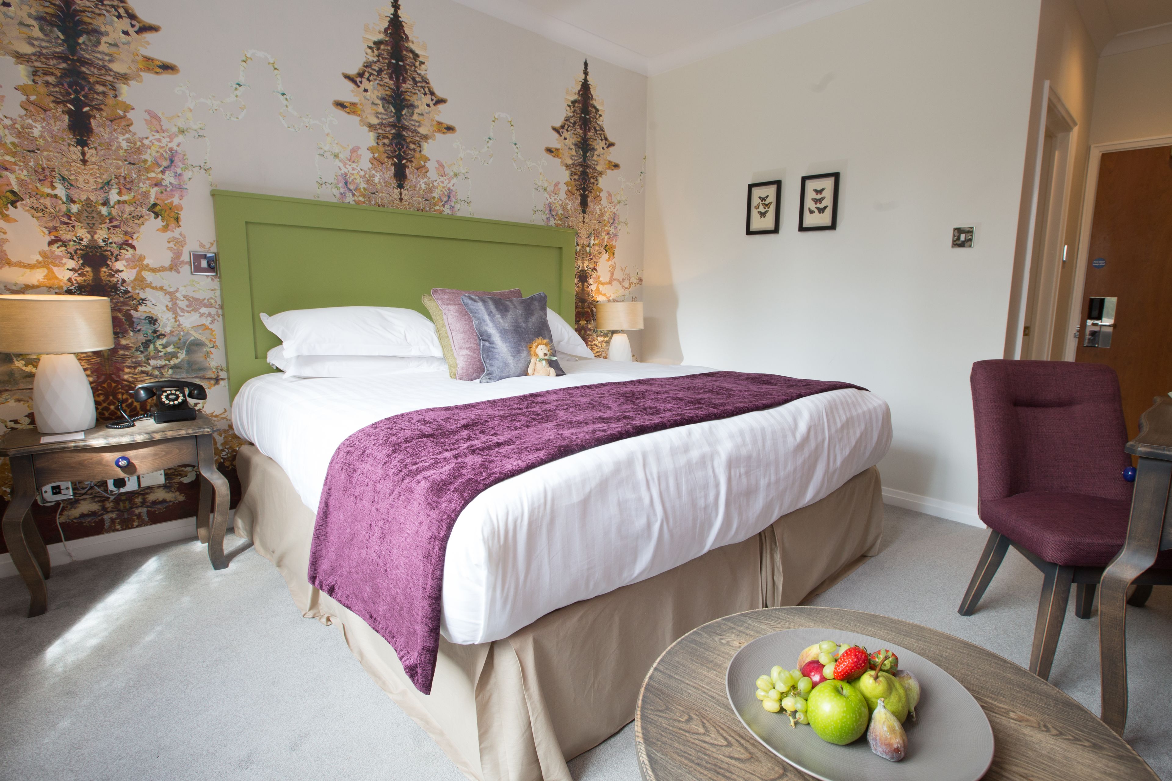 A hotel room with a green headboard, a bed with white linens and a purple throw, a round wooden table with fruit, and a purple armchair.