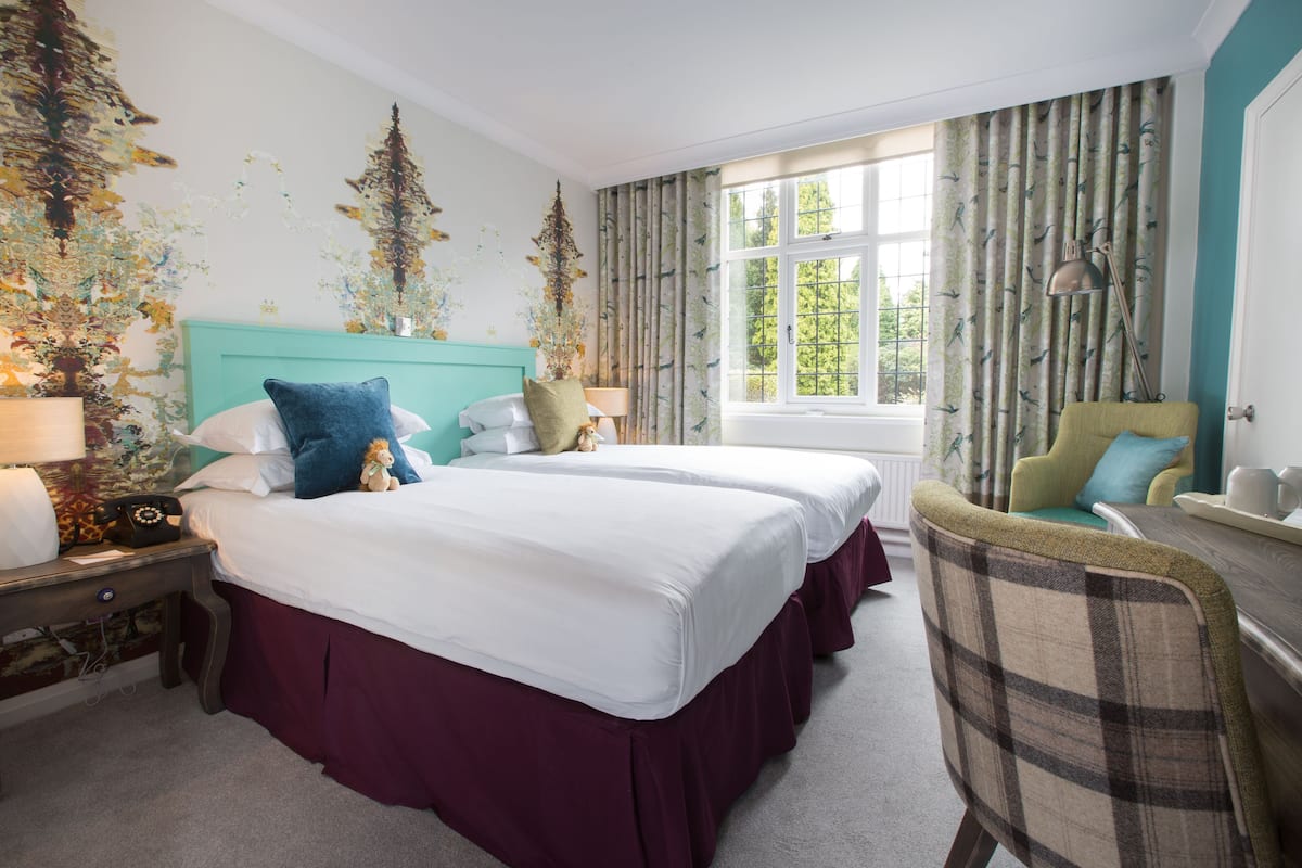 A hotel room with two beds, a chair, a nightstand, and a window with curtains.