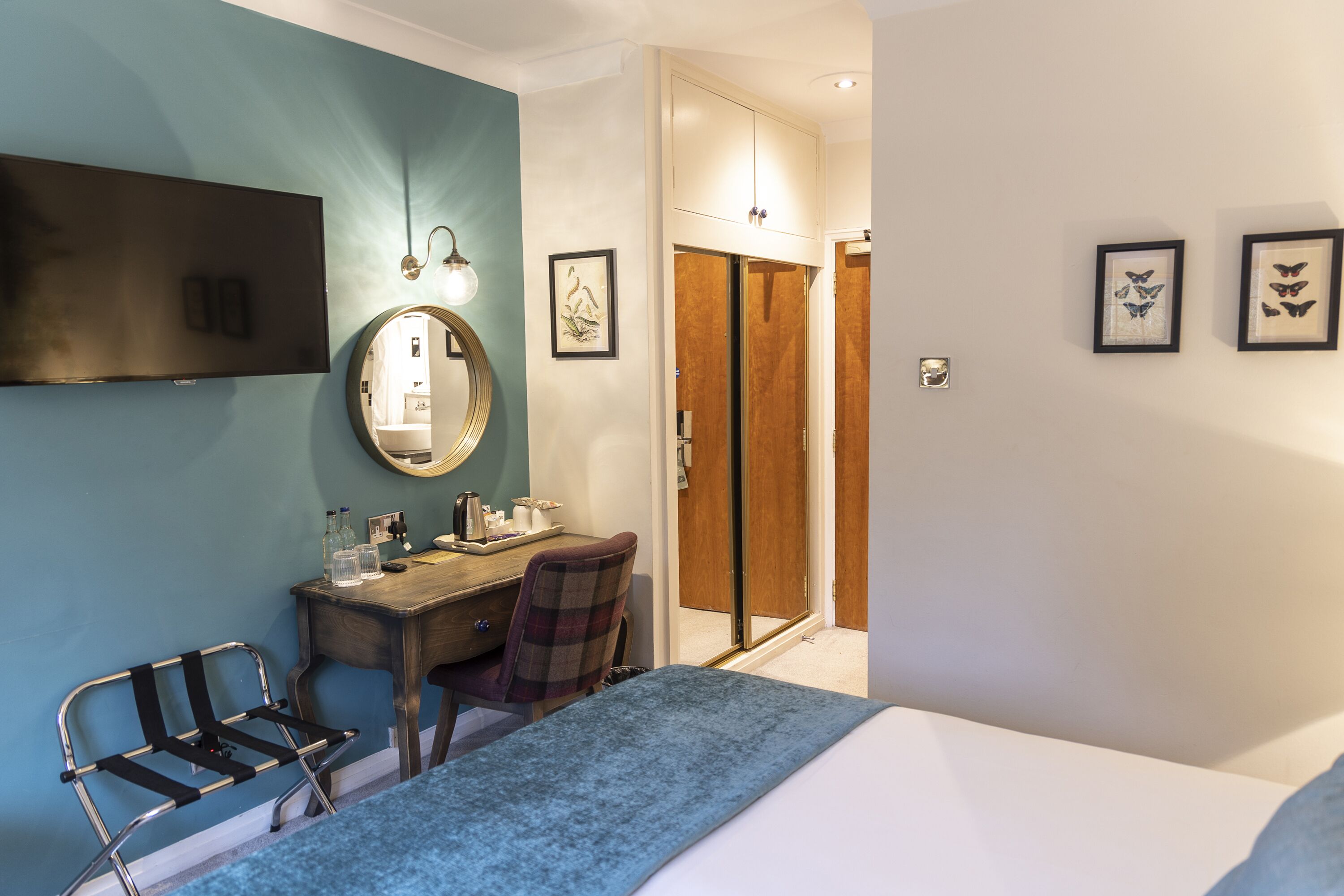 Een hotelkamer met een bed, een bureau met stoel, een televisie, een spiegel en beelden in lijsten aan de muur.
