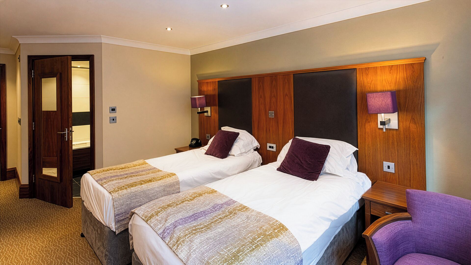 A hotel room with two beds, a wooden headboard, a bedside table with a lamp, and a chair.